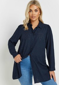 Camicia blu scuro con maniche lunghe, apertura frontale con bottoni e motivo in occhietti. Presenta un collo e una vestibilità rilassata, realizzata in tessuto leggero.