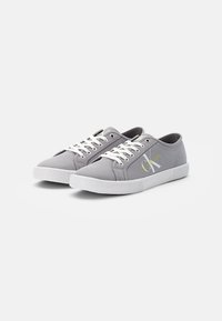 Graue Leinwand-Sneaker mit weißen Schnürsenkeln, gummierter Sohle und strukturierten Details. Seitlich gelbes Logo. Klassisches Low-Top-Design.