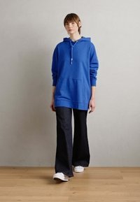 Blauer Hoodie mit einer vorderen Tasche, Kordelzugkapuze und gerippten Bündchen, kombiniert mit dunklen Schlagjeans und weißen Turnschuhen auf einem Holzboden.
