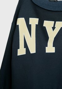 Sweat-shirt noir avec de grandes lettres crème "NY" imprimées sur le devant, montrant la partie supérieure du torse et une partie du col.