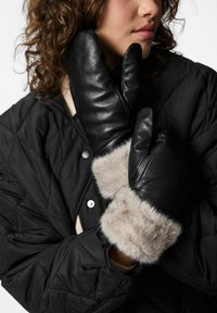 Juodos odos pirštinės su minkštu pilku kailiu ant rankogalių, pasižyminčios lygiu paviršiumi ir prigludusiu dizainu. Dėvimos ant juodos siūtos striukės.