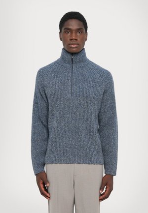 Homme portant un pull en tricot bleu à col montant et un pantalon gris clair, se tenant devant un fond blanc uni.