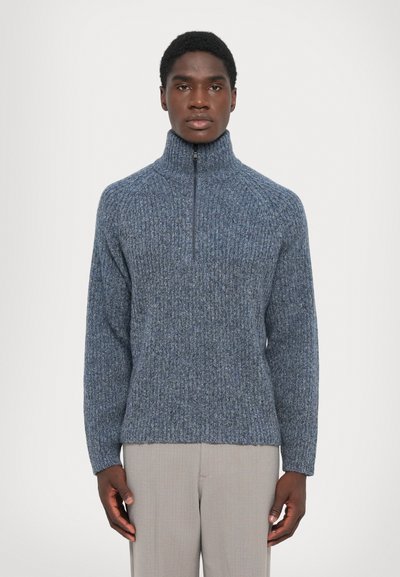 Mann in einem blauen, hoch geschlossenem Zip-Pullover und hellgrauen Hosen, der vor einem schlichten weißen Hintergrund steht.