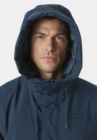 Giacca a cappuccio blu navy con chiusura a zip, cordini regolabili e logo in gomma sul petto. Texture liscia, design funzionale e forma aderente.