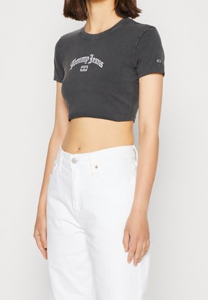 Frau trägt ein eng anliegendes, kurzes schwarzes Tommy Jeans T-Shirt und hoch taillierte weiße Hose, steht mit entspannten Armen an den Seiten.