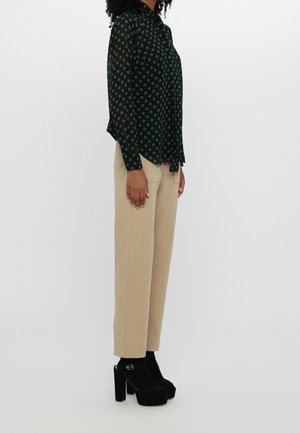 Vrouw draagt een zwarte blouse met groene stippen, beige rechte broek en zwarte plateaus met hakken, staand in profiel op een witte achtergrond.