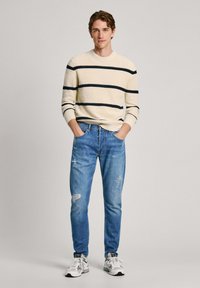 Vaaleanbeige neulepaita, jossa on tummansiniset raidat, yhdistettynä kuluneisiin vaaleansinisiin farkkuihin ja valkoisiin lenkkikenkiin. Malli poseeraa kädet taskuissa.