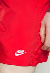 Červené šortky Nike vyrobené z ľahkej tkaniny s bielym vyšívaným logom na ľavej strane a viditeľným šitím pozdĺž švov.