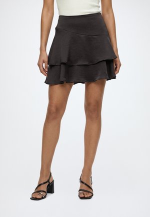 Femme portant une mini-jupe noire à volants avec une taille ajustée et des sandales à talons noires avec des brides croisées.