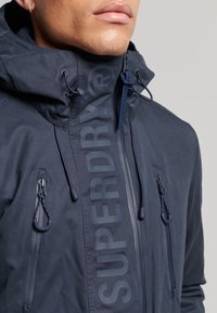 Zbliżenie osoby w granatowej kurtce z wysokim kołnierzem, regulowanymi sznurkami, zamykanymi kieszeniami na piersi oraz logo „SUPERDRY” na klapie zamka.