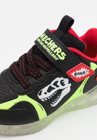 Sapatilhas infantis pretas e vermelhas com detalhes em verde-neon, gráfico de crânio de dinossauro, patch "ROAR!", tira de Velcro rotulada "Skechers Resistente à Água".
