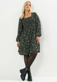 Musta kukkakuvioinen mekko, jossa on neliskulmainen kaulus, pitkät puffihihat, kerätty vartalo ja levenevä helma. Suunniteltu mustien nauhakengien kanssa.