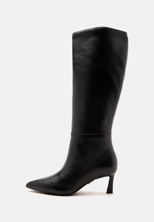 Botte en cuir noire montant jusqu'au genou avec bout pointu et talon fin de hauteur moyenne, vue de profil sur un fond uni.