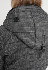 Grijze gewatteerde jas met een capuchon, met een gestructureerd, geruit patroon en een ronde zwarte knop dichtbij de kraag. Verstelbare trekker aan de achterkant.