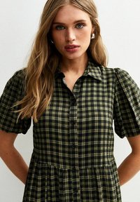 Robe chemise à carreaux verts avec col, manches courtes bouffantes, devant à boutons et taille froncée. Le tissu a une apparence légère et texturée.
