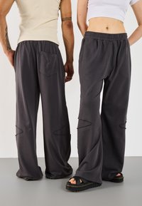 Two people wearing loose dark gray pants, one with a beige sleeveless top and one with a white cropped top and black sandals against a plain background.