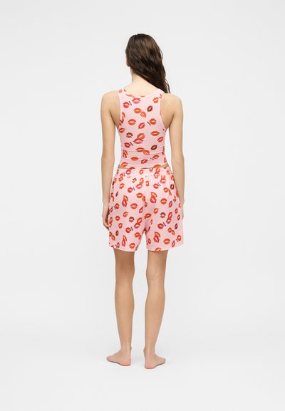 Γυναίκα όρθια, ξυπόλυτη, γυρισμένη πλάτη, φοράει ανοιχτό ροζ μπλουζάκι και σορτς με κόκκινα και πορτοκαλί σχέδια από τύπωμα χειλιών.