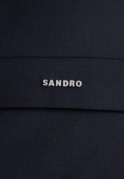 Tessuto nero con pieghe orizzontali presenta un logo argento con la dicitura "SANDRO" in evidenza. La texture è liscia e uniforme.