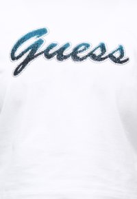 Λευκό ύφασμα με μπλε και μαύρο στρας που σχηματίζουν το γράμμα "Guess" περιγραμμένο με μικρές ασημένιες χάντρες.