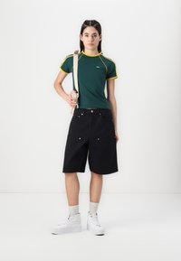 Grønn T-skjorte med gule detaljer, svarte shorts og hvite høy-top sneakers. Modell som holder en beige veske. Enkelt design og uformell passform.