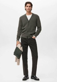 Sivý cardigan s V-krkom a piatimi gombíkmi, skombinovaný s tmavými džínami. Model drží sveter so zeleno-krémovými pruhmi. Čierne topánky.