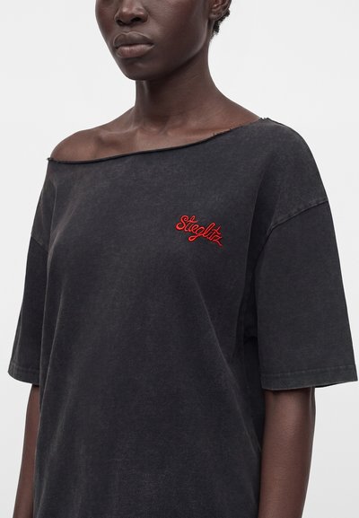 Γυναίκα με μαύρο χαλαρό T-shirt με ακατέργαστη λαιμόκοψη και κόκκινο καλλιγραφικό λογότυπο "Siegltz" στο πάνω αριστερό μέρος του στήθους.