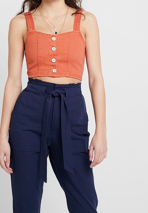 Top corto color ruggine con spalline larghe e dettaglio di bottoni frontali, abbinato a pantaloni alta vita blu navy con cintura legata e tasche.