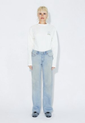 Weißes Langarmshirt mit einer Grafik auf der Brust, kombiniert mit hellblauen Jeans mit weitem Bein. Das Model steht vor einem schlichten Hintergrund und trägt schwarze Schuhe.