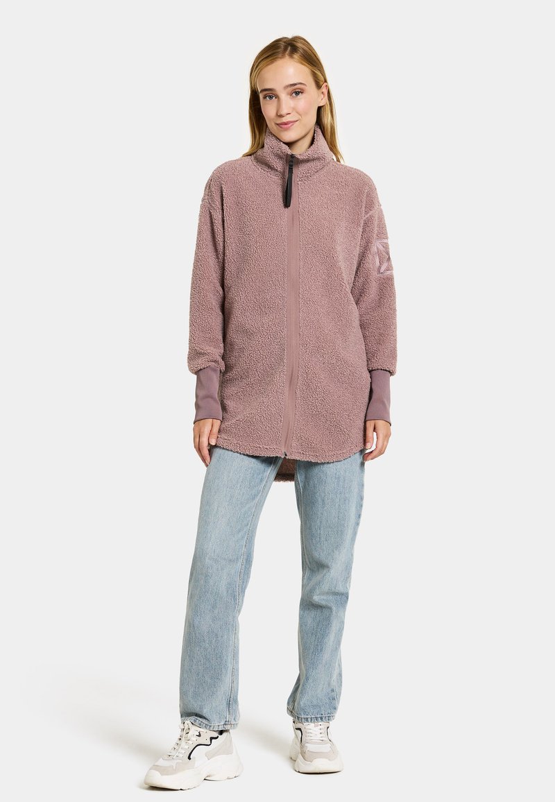 Frau, die eine mauvefarbene Fleecejacke mit langen Bündchen, hellblaue Jeans und weiße, klobige Sneakers trägt, steht vor einem einfarbigen, hellen Hintergrund.