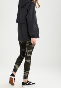 Musta kevyt huppari leveällä mallilla, yhdistettynä vihreän ja ruskean sävyisiin tiukkoihin maastokuvioisiin legginsseihin sekä mustiin tennareihin, joissa on valkoiset pohjat.