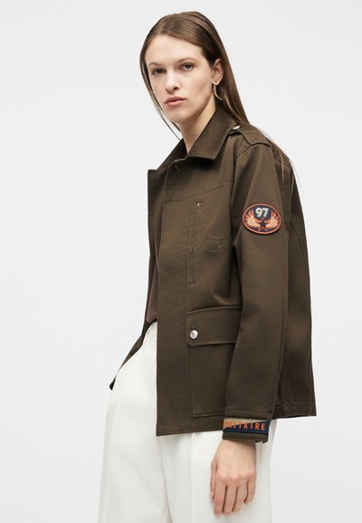 Γυναίκα που φορά καφέ μπουφάν με επιθέματα "97" και "Voltaire" στο μανίκι, συνδυασμένο με άσπρο παντελόνι, στέκεται μπροστά σε απλό φόντο.