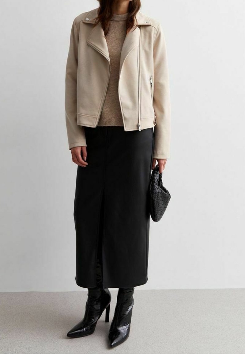 Béžová umělá kožená motorkářská bunda, hnědý pletený svetr, černá kožená sukně s bočním rozparkem a černé boty na vysokém podpatku s špičatou špičkou. Černá kabelka.