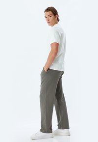 Vit kortärmad skjorta kombinerad med grå texturerade byxor och vita sneakers, stående framför en neutral bakgrund med händerna i fickorna.