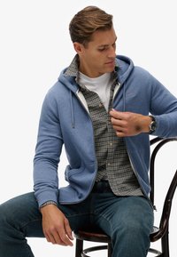Sweat à capuche bleu avec une texture douce, poches avant, et cordon de serrage décoratif, superposé à une chemise à carreaux, porté avec un jean sombre.