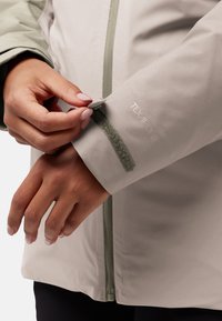 Lichtgrijze waterdichte jas met een gladde textuur, een verstelbare klittenbandmanchet en een voorrits met een groene acccent.