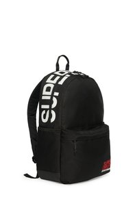 Zaino in tessuto nero con spallacci regolabili, forma curva, tasca frontale con zip e audace logo bianco "SUPERDRY" sul lato, dettagli rossi.