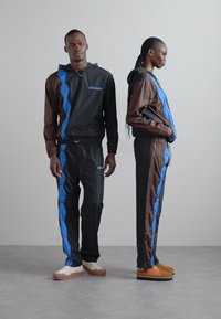 Svarta och bruna träningsdräkter med blå sidosträck, bestående av en zip-up hoodie och byxor med elastisk midja, tillverkade av ett mjukt och lätt tyg.