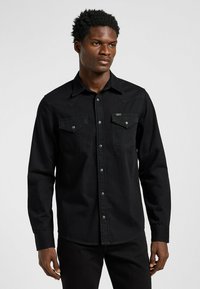 Camisa negra de manga larga de mezclilla, con dos bolsillos en el pecho, botones a presión y un cuello puntiagudo con costuras en contraste.