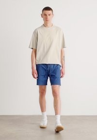 T-shirt oversize beige clair, shorts bleus, chaussettes blanches et baskets ; tenue décontractée avec une coupe ample et des couleurs unies. Design minimaliste.