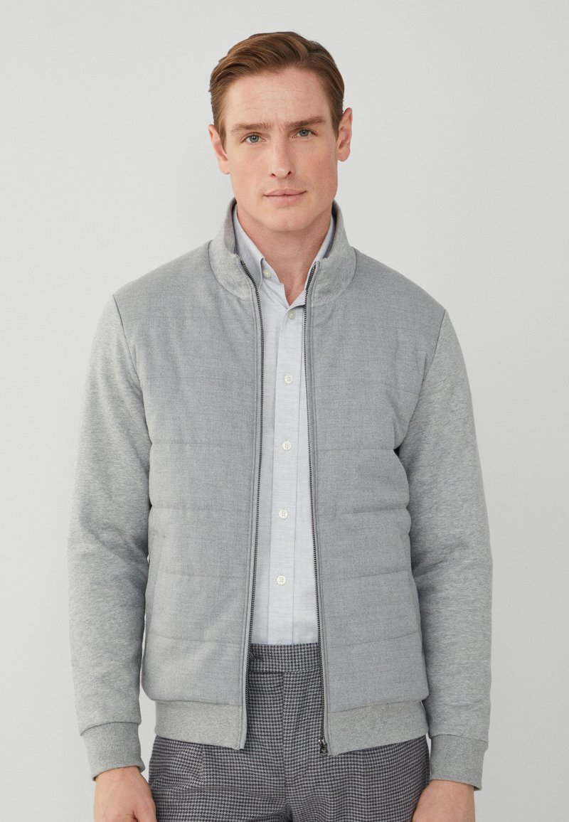 Graue Zip-Jacke mit hohem Kragen, gestepptem Muster und kontrastierenden weichen Ärmeln. Tragen über einem hellen Hemd und karierter Hose.