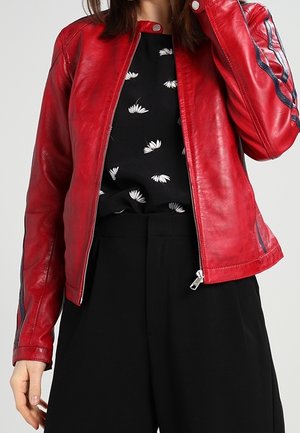 Femme portant une veste en cuir rouge partiellement zippée avec une chemise fleurie noire et blanche et un pantalon noir taille haute.