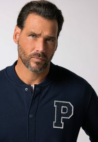 Marinblå sweatshirt med rund halsringning och två tryckknappar. Har en strukturerad vit "P"-logotyp på vänster bröst. Slätt tyg, avslappnad design.