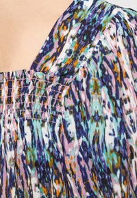 Färgglad mönstrad blus med smockad halsringning. Har en blandning av lila, svart, turkos och orange i ett flytande, lätt tyg.