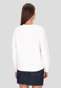 Sudadera blanca de forro polar con puños y dobladillo acanalados, con cuello redondo y mangas raglán. Combinada con una falda vaquera. Vista trasera.
