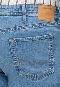 Jeans de mezclilla azul claro con un bolsillo trasero que presenta detalles de costura y una etiqueta de cuero con "JACK & JONES". Textura suave, corte clásico.