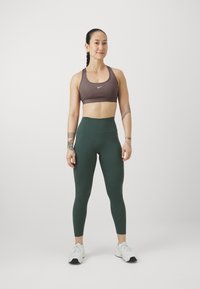 Soutien-gorge de sport marron avec un logo blanc, leggings taille haute verts et chaussures de sport blanches, debout sur un fond uni.