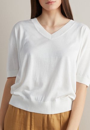 Kobieta w białym swetrze z krótkim rękawem i dekoltem w serek, włożonym do wysokich brązowych spodni, widoczna od ramion do bioder.