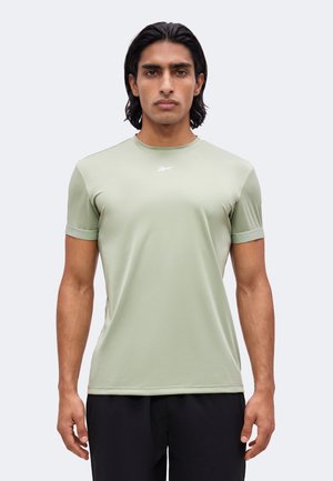 Uomo con capelli neri di media lunghezza che indossa una maglietta sportiva verde chiaro a maniche corte e pantaloni neri, rivolto verso l'obiettivo con espressione neutra.