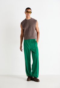Hombre vistiendo una camiseta sin mangas marrón de punto, pantalones verde de pierna ancha, zapatos marrones y gafas de sol con tintes amarillos, de pie contra un fondo blanco.