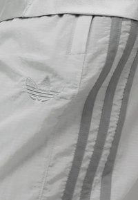 Šviesiai pilki sportiniai šortai su siuvinėtu logotipu, vertikaliomis juostelėmis šiek tiek tamsesnėje atspalvyje ir tekstūruota medžiaga.